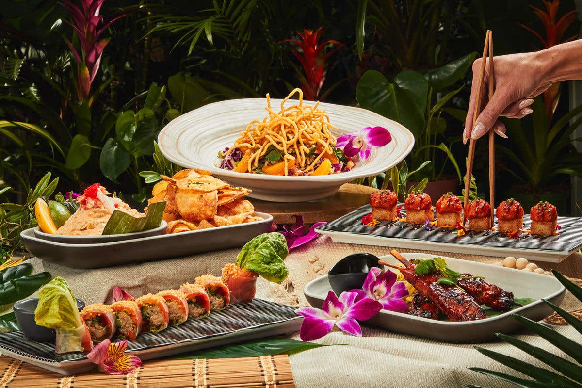 An assortment of Asian-inspired dishes featuring sushi, salad, skewers, chips, and bite-sized morsels on a table, accented with orchids and greenery.