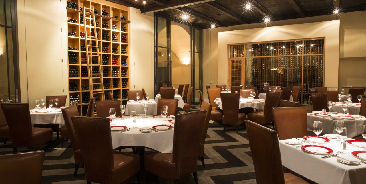 Elegant restaurant interior with leather chairs, round tables with white cloths, and shelves filled with wine bottles on walls.