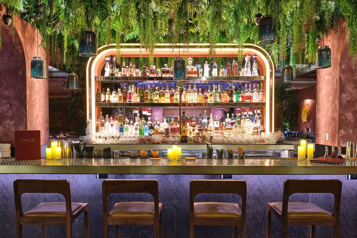 A dimly lit bar with lush hanging plants above, a variety of bottles on shelves, and candles on the counter with four bar stools.