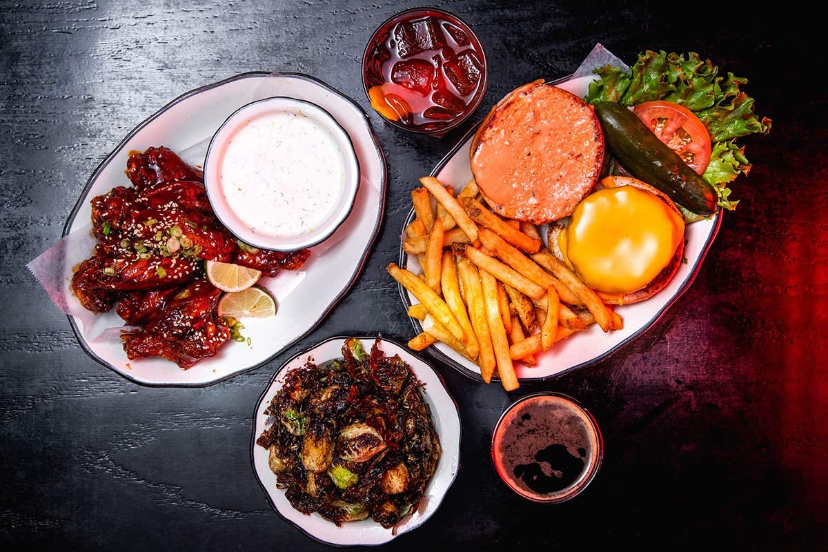 Plates of BBQ wings, fries with a cheeseburger, and roasted Brussels sprouts, with drinks on a black table.