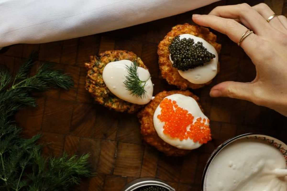 Three latkes with cream and roe toppings on a wooden board, with dill and a bowl of sauce. A hand reaches for one latkes.