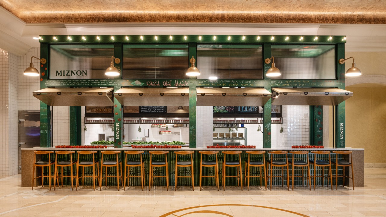 A restaurant counter with wooden bar stools, green accents, and a sign reading "Miznon" above.