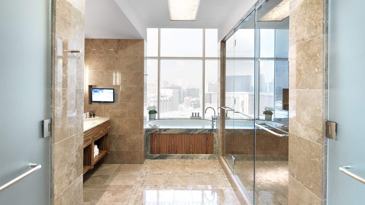 Luxurious bathroom with marble floors, walk-in shower, bathtub, large window view of a cityscape, and mounted TV.