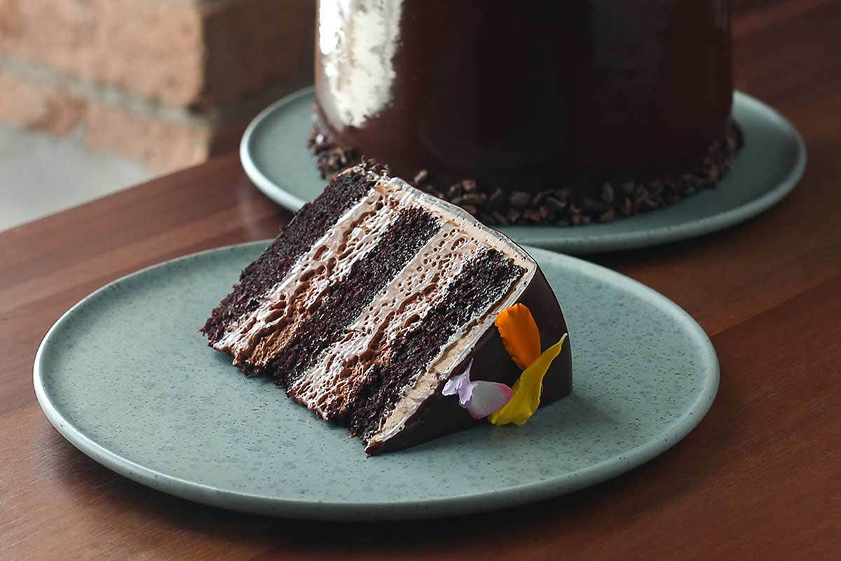 A slice of chocolate layer cake with brown frosting, garnished with edible flowers, on a gray plate.