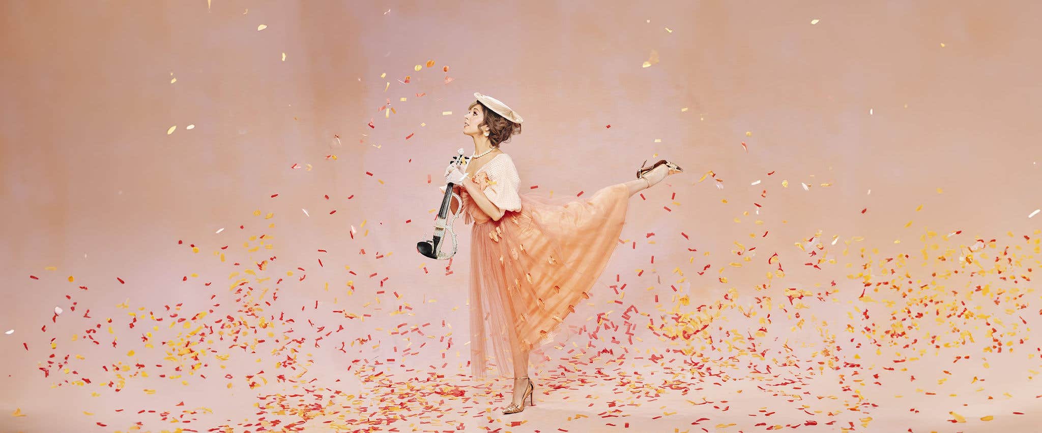 Lindsey Stirling posing with an electric violin standing on one leg whilst against a light orange backdrop with confetti