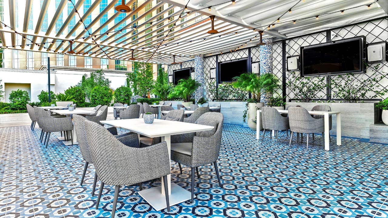 Outdoor patio dining area with grey woven chairs, white tables, blue patterned tiles, greenery, and large TVs.