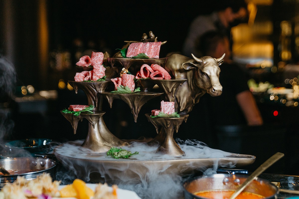 Cold cuts displayed on a multi-tiered, ornate metal stand resembling a bull, with a smoky effect at the base.