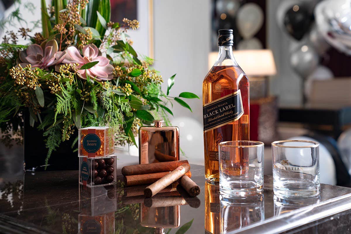 Bottle of Black Label whiskey, cigars, glasses, chocolates, and a copper canister on a table beside a floral arrangement.