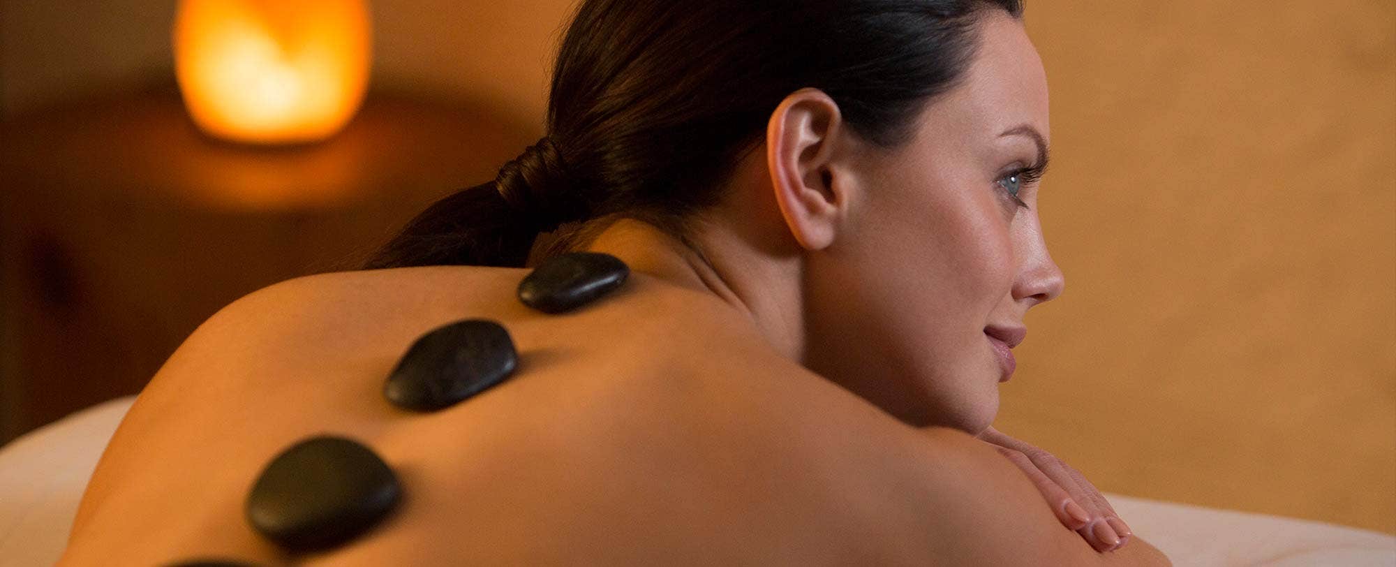 A woman receives a hot stone massage while lying on a massage table, with smooth stones placed on her back.