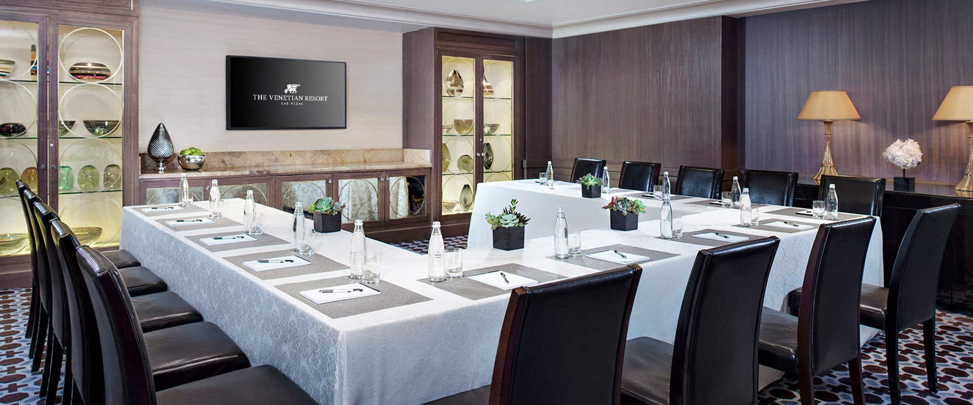 A room with table setup in a horseshoe shape that seats 12 people and a TV centred on the wall.