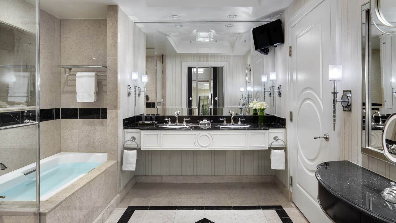 Luxurious bathroom with a double-sink vanity, bathtub with towel, large mirror, wall-mounted TV, and modern lighting.