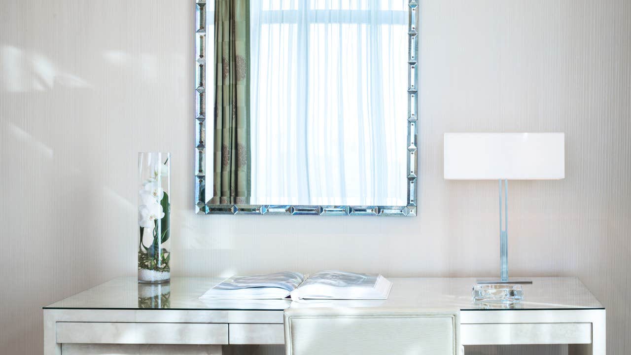 A minimalist desk setup with a glass vase, an open book, a modern lamp, and a large framed mirror reflecting a window.