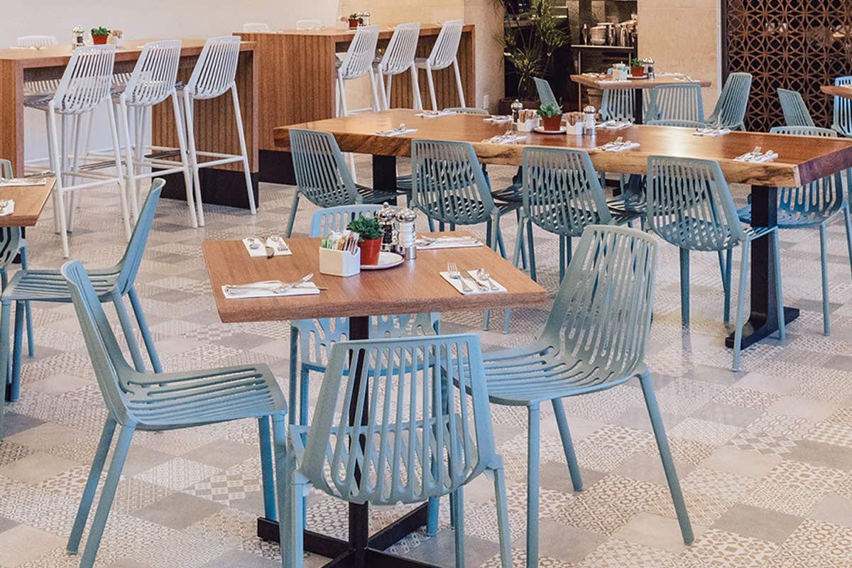 Modern café interior with wooden tables, blue and white chairs, set with cutlery and small potted plants, tiled floor.