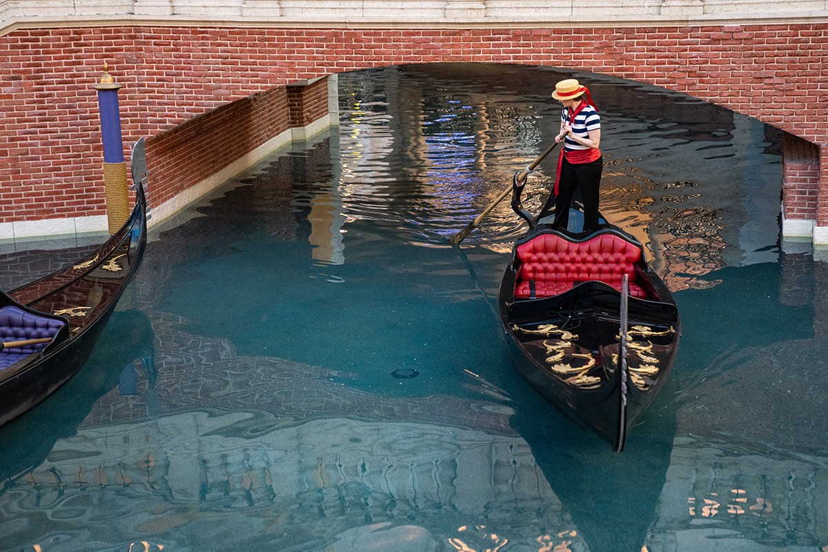 A gondolier navigates a black gondola under a brick bridge in a canal with two gondolas visible.