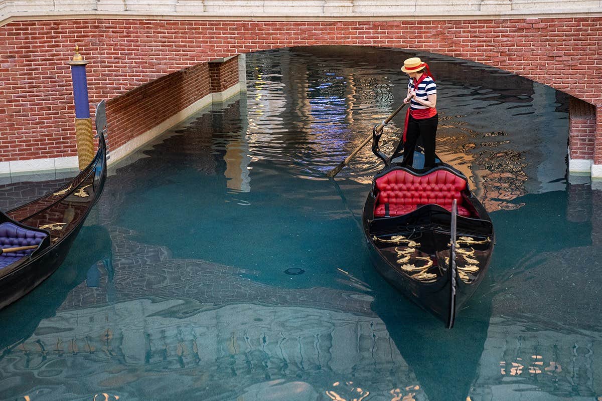 A gondolier navigates a black gondola under a brick bridge in a canal with two gondolas visible.