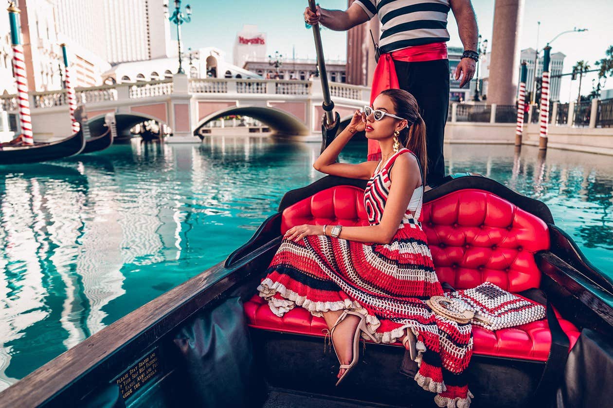 Woman in a red dress and sunglasses sits in a gondola on a canal with Venetian-style architecture in the background.