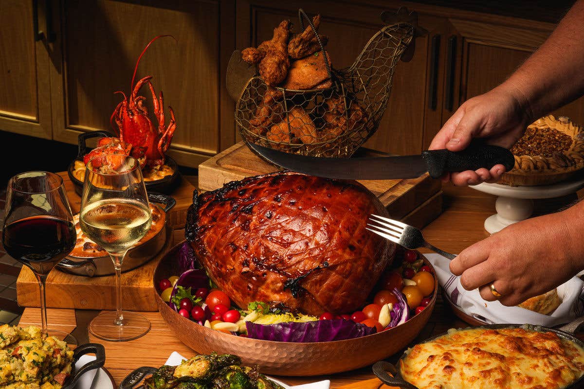 A dinner table displaying a glazed baked ham being sliced, alongside wine glasses, lobster, and various side dishes.