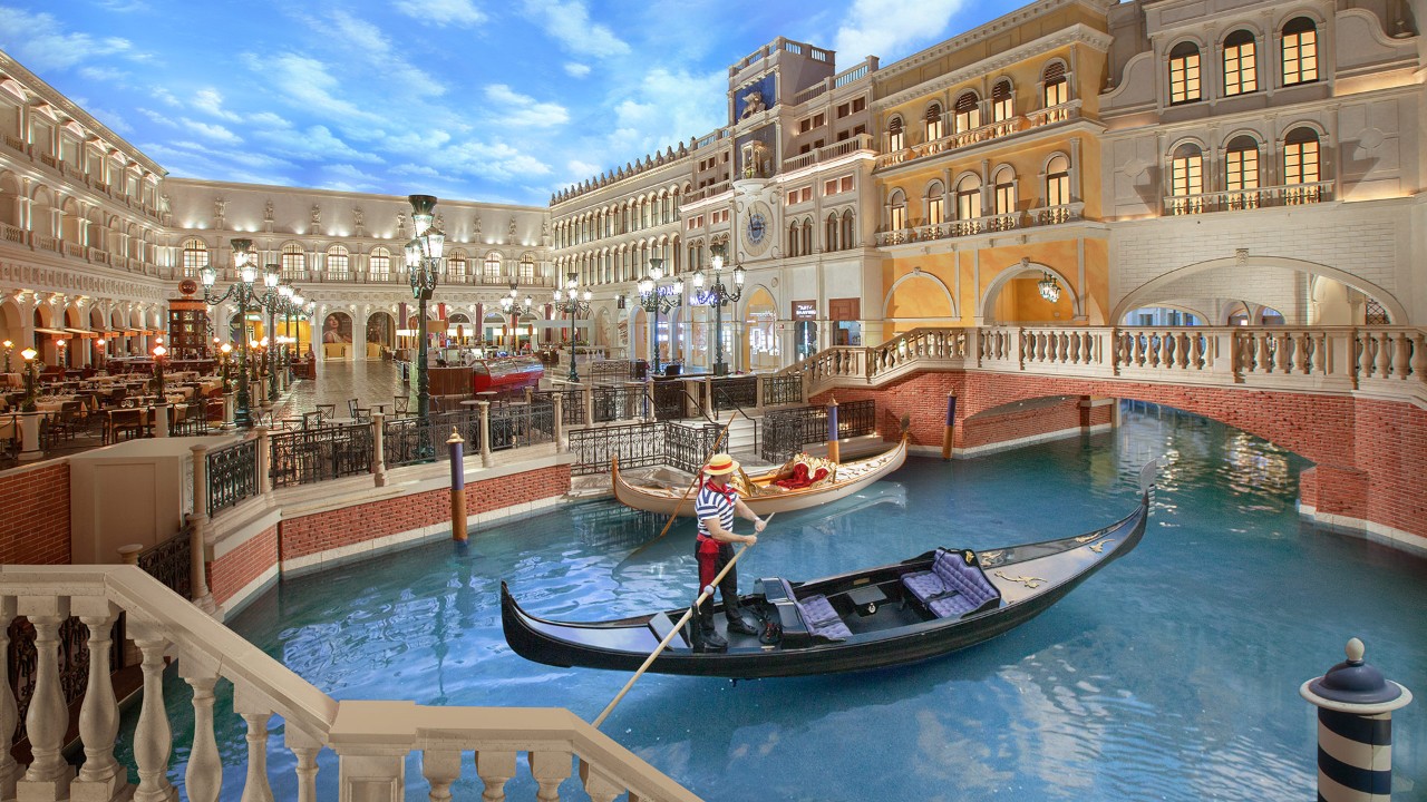 A gondola navigates an indoor canal surrounded by Italian-themed buildings with bright, clear skies above.