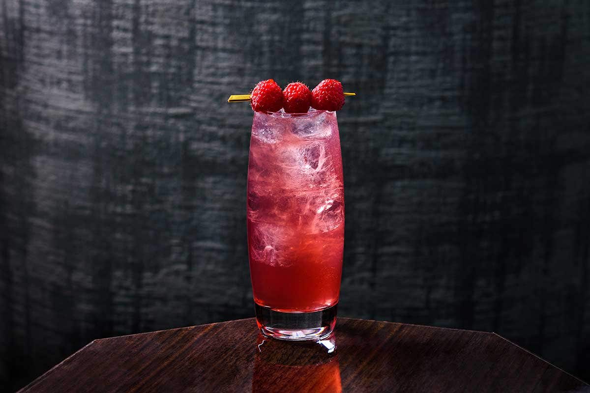 A tall glass of red cocktail on a wooden table, garnished with three raspberries on a skewer.