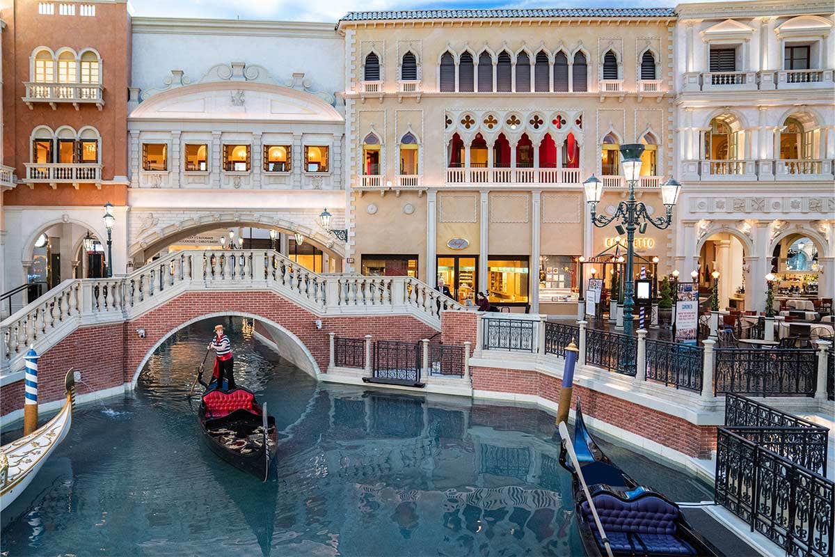 Indoor scene of a Venetian canal with gondolas and ornate buildings, featuring a bridge and shops in the background.