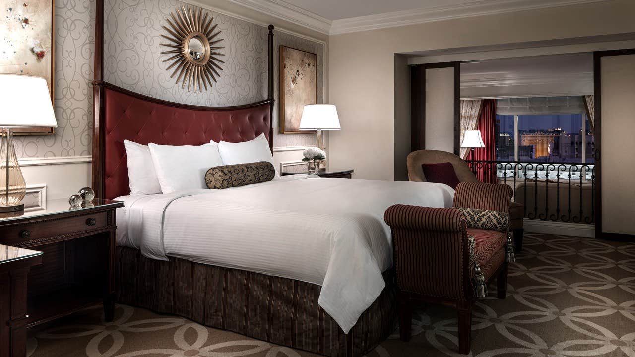 Luxurious hotel room with a large bed, two side tables with lamps, upholstered chairs, and a city view through sliding doors.