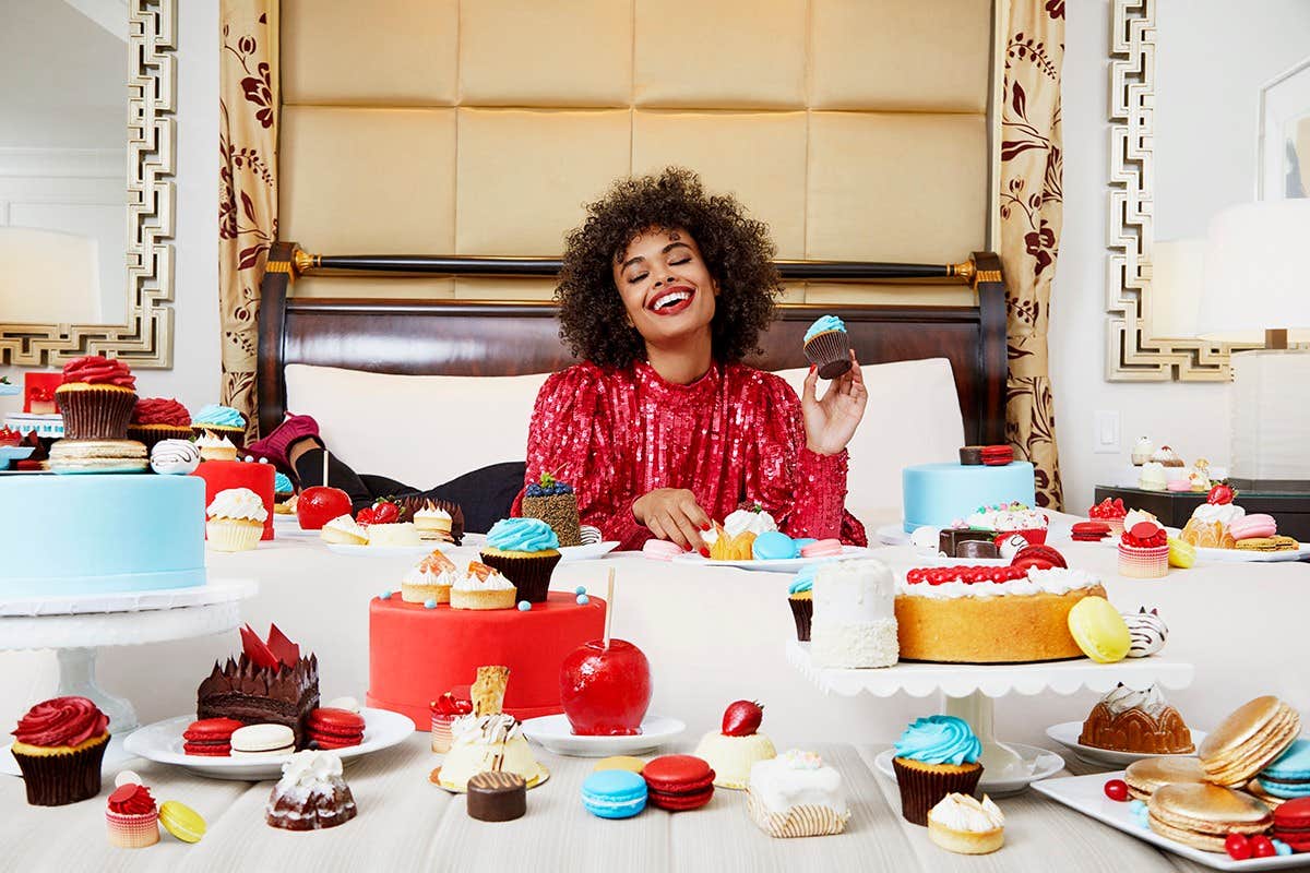 A joyful person in a red sequin top surrounded by a variety of colorful desserts on a bed in a stylish room.