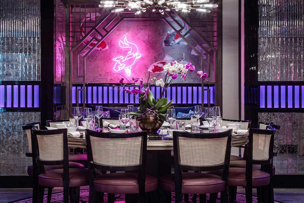 Elegant dining area with round table, set with glassware and a floral centerpiece, neon pink sign in the background.