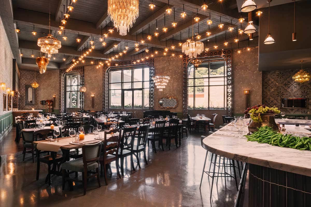 Elegant restaurant interior with chandeliers, large windows, and a mix of tables and a bar area with stools.