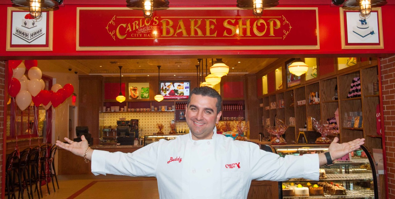 A person stands with arms open in front of "Carlos Bake Shop," wearing a white shirt that says "Buddy.