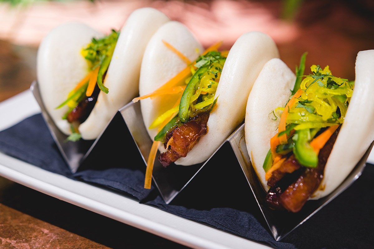 Three steamed bao buns filled with colorful vegetables and meat, served on a black napkin on a rectangular white plate.