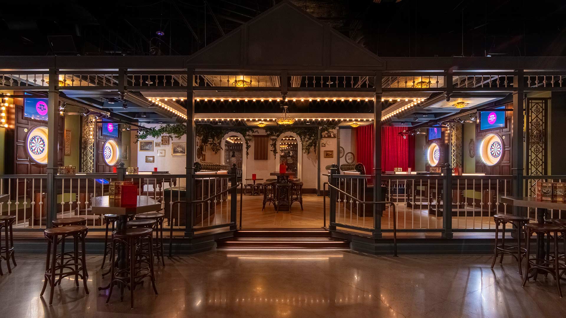 A cozy bar with dartboards, wooden tables, and stools, illuminated by warm lighting and string lights.