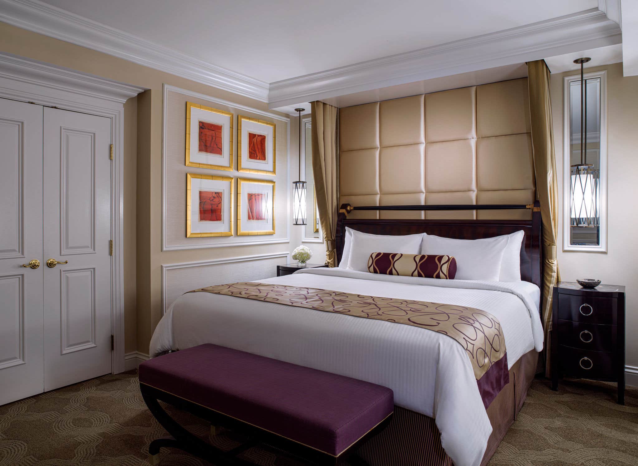 Elegant hotel room with a king-sized bed, purple bench, framed artwork, bedside lamps, and decorative pillows.