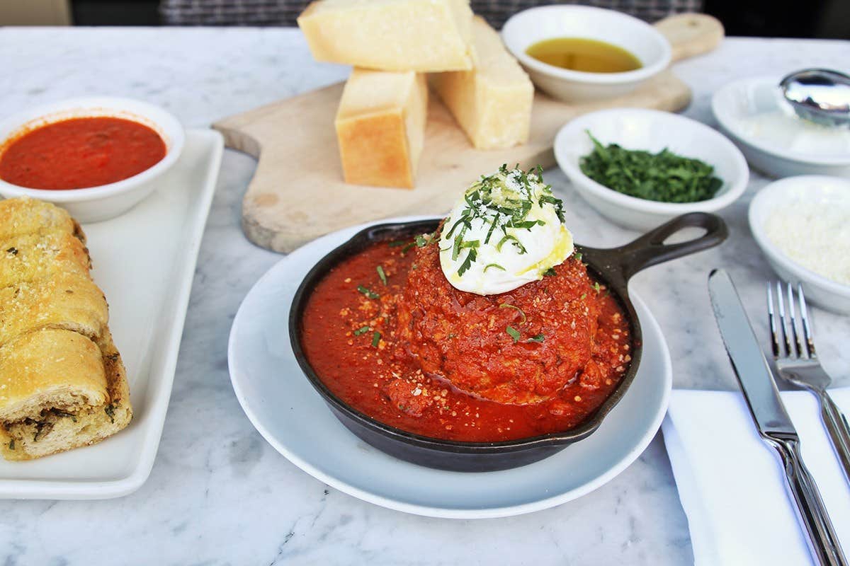 A skillet with a large meatball topped with ricotta and basil, surrounded by bread, sauce, and condiments on a marble table.