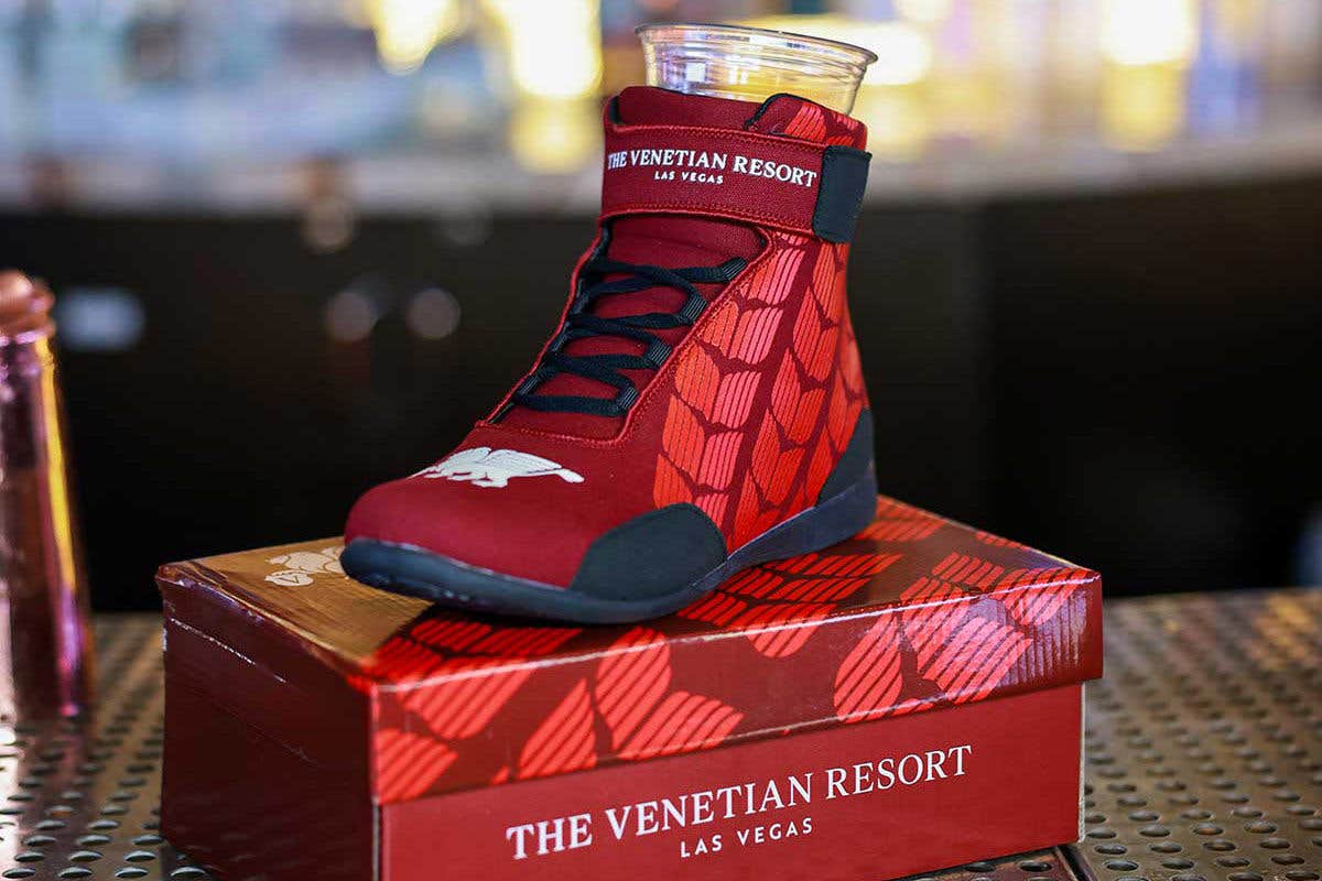 Red racing shoe with The Venetian Resort Las Vegas logo with a plastic cup sitting inside of it, resting on a shoe box on a bar