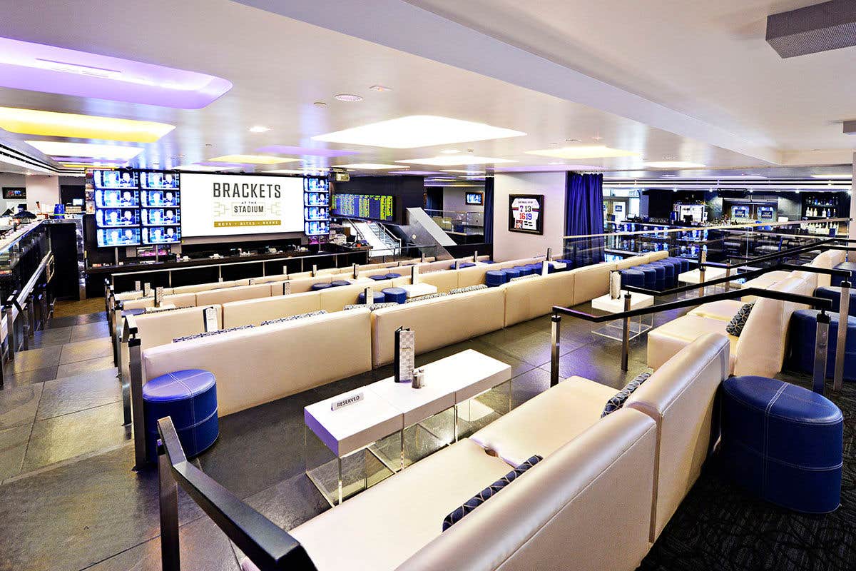 Upscale sports bar with cream couches, blue ottomans, and large screen displaying "Brackets At The Stadium."