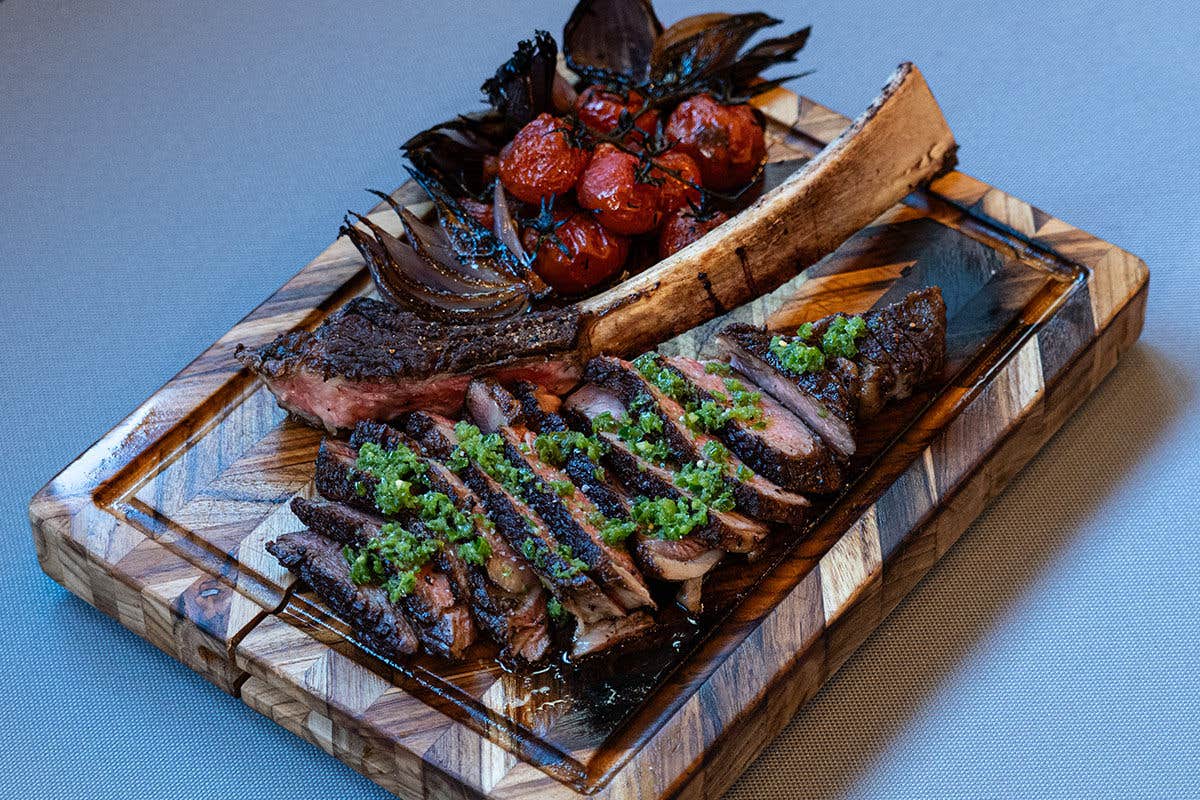 A bone-in ribeye steak sliced and presented with a green sauce on top, next to the bone and some blistered tomatoes on a wooden cutting board