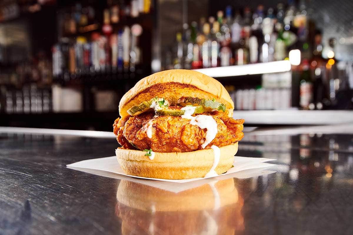 Close-up of a spicy fried chicken sandwich with pickles, jalapeños, and white sauce on a shiny counter. Bar in the background.