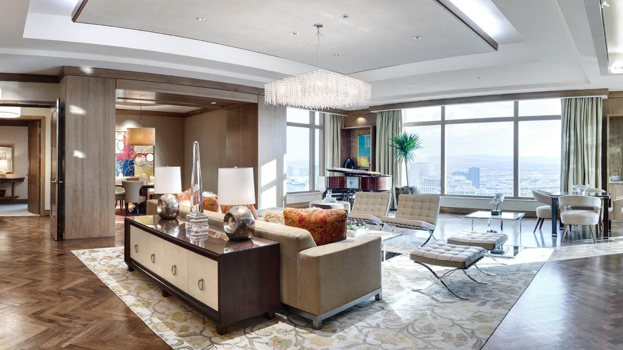 Spacious modern living room with large windows, a chandelier, cozy seating, and a grand piano overlooking a cityscape.