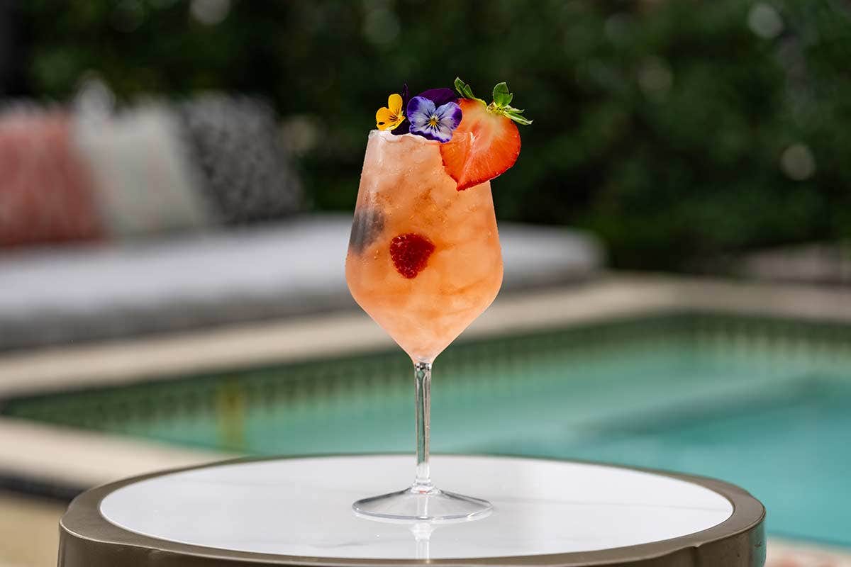 A refreshing orange-pink cocktail garnished with a flower and strawberry slice, placed on a table by a pool.