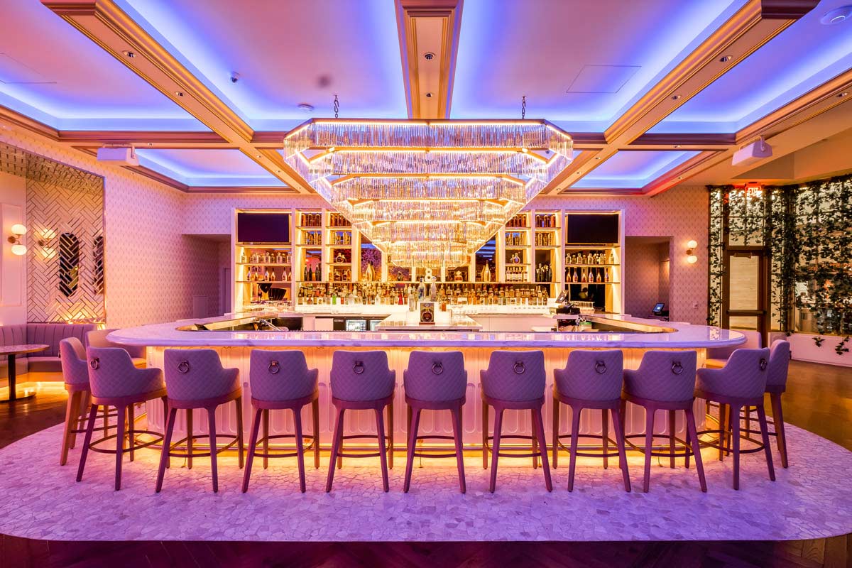 Luxurious bar with a central chandelier, surrounded by upholstered bar stools and illuminated by ambient purple lighting.