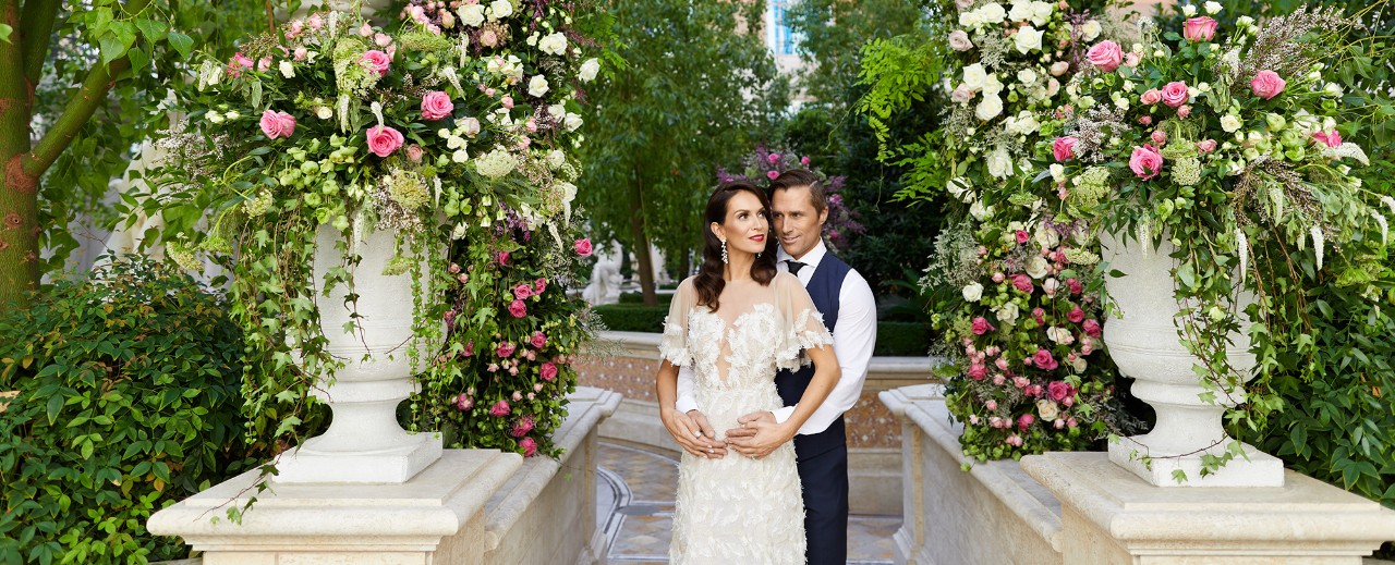 A couple in wedding attire stand embraced, surrounded by lush foliage and vibrant floral arrangements.