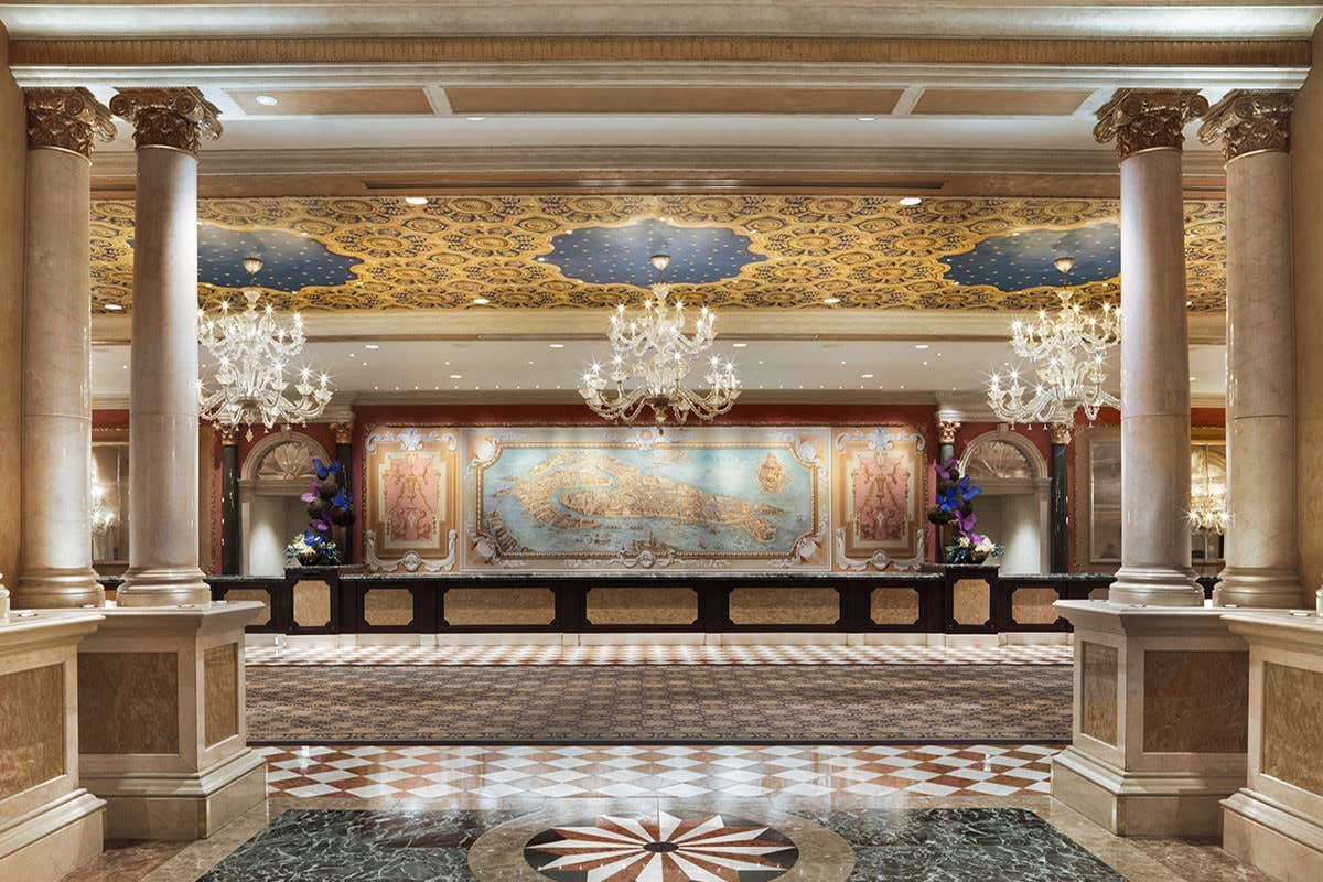 Elegant hotel lobby with ornate chandeliers, decorative ceiling, marble columns, and a detailed wall map behind the reception.