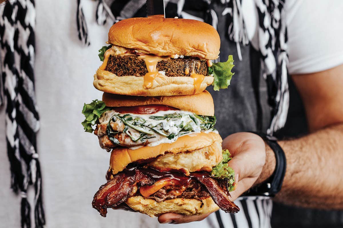 Person holding a stack of three burgers with various toppings, including lettuce, bacon, and a creamy sauce.