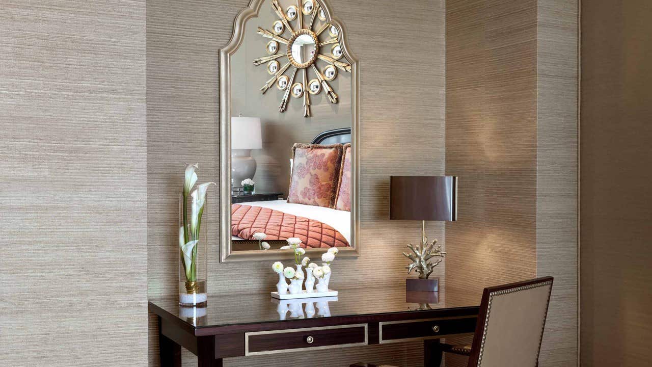 A stylish vanity with a mirror, decorative items, a lamp, and a chair, reflecting a bed with pillows in a tastefully decorated room.