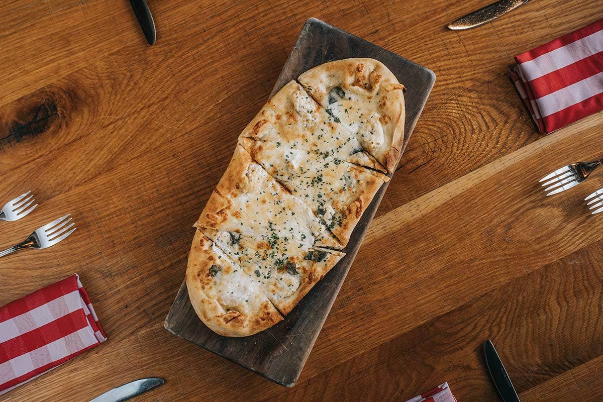 Flatbread pizza on a wooden board, surrounded by silverware and red checkered napkins on a wooden table.