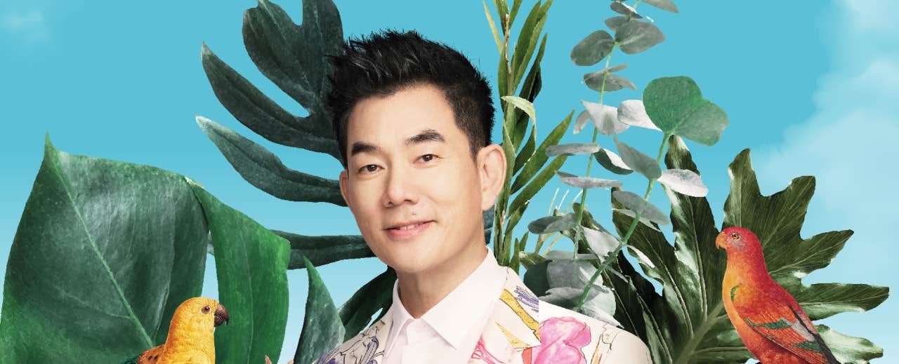 A man in a floral blazer stands among lush greenery with birds perched on the plants around him.