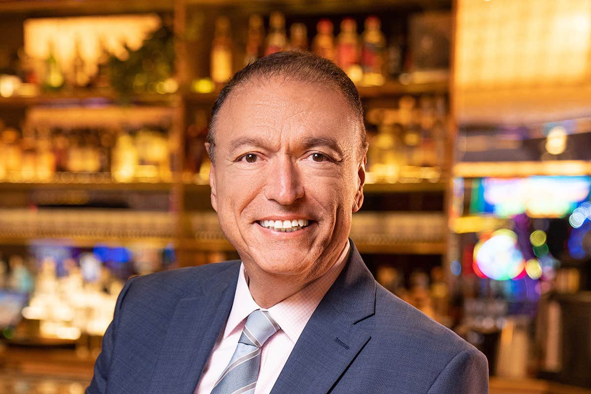 A smiling man in a suit and tie stands in a warmly lit bar with shelves of bottles in the background.