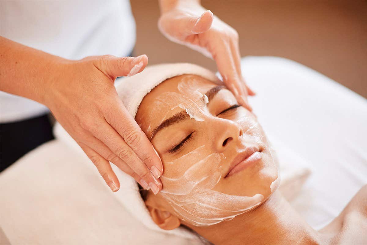 A person receiving a facial treatment with a creamy mask applied to their face, eyes closed, and a towel on their head.