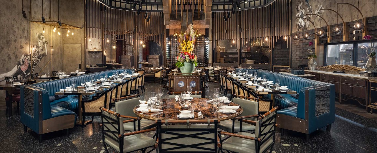 Elegant restaurant interior with blue upholstered booths, round tables with floral centerpieces, and rustic decor elements.