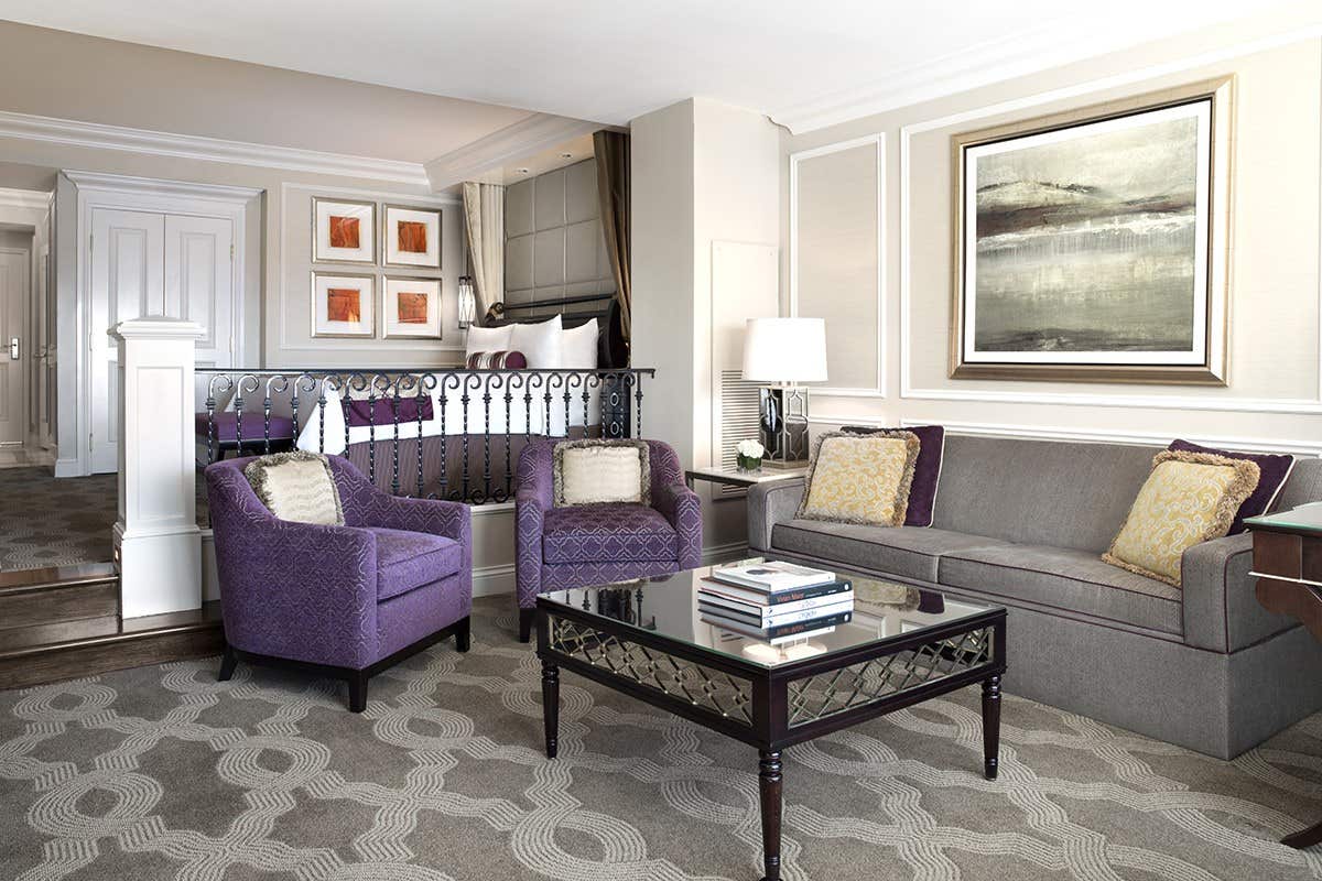 Stylish hotel room with a gray sofa, two purple armchairs, a glass coffee table, and framed artworks on the walls.